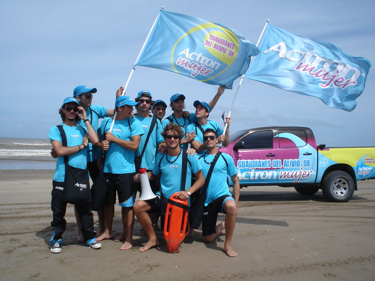 Equipo con remeras y gorras celestes de Actron Mujer en una playa, junto a banderas de la marca