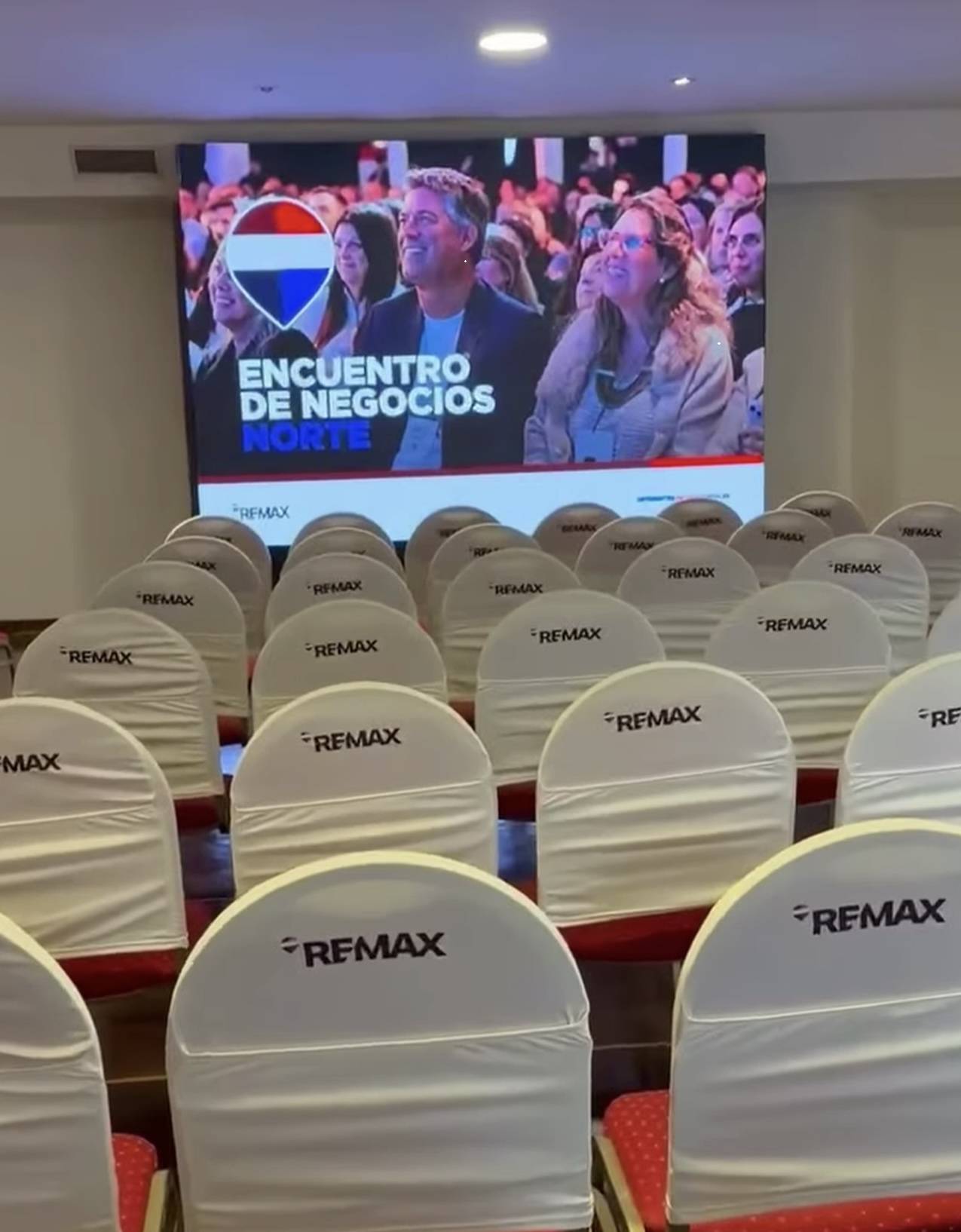 Sala del Encuentro de Negocios Norte con fundas de sillas RE/MAX y pantalla del evento